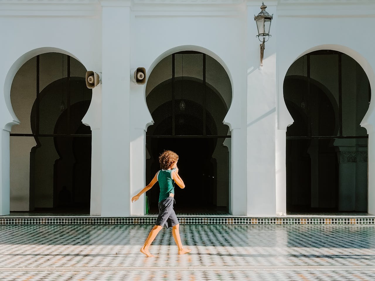 A child strolls in a sunlit Moroccan courtyard featuring traditional architecture with arches.